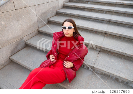 High fashion photo of a young brunette woman in red glossy down jacket, and red pants. Urban street fashion. High fashion photo of a young brunette woman in red glossy down jacket, and red pants. Urban street fashion. 126357696