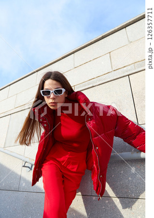 High fashion photo of a young brunette woman in red glossy down jacket, and red pants. Urban street fashion. 126357711