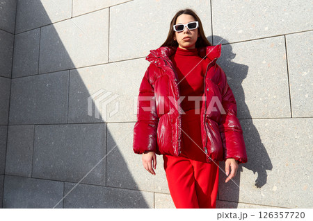High fashion photo of a young brunette woman in red glossy down jacket, and red pants. Urban street fashion. High fashion photo of a young brunette woman in red glossy down jacket, and red pants. Urban street fashion. 126357720