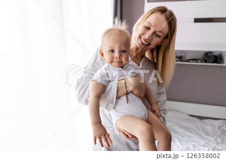 Smiling beautiful mother holding baby in bright white room, dressed in white, natural daylight, happy family, copy space on background, lifestyle and parenthood 126358002