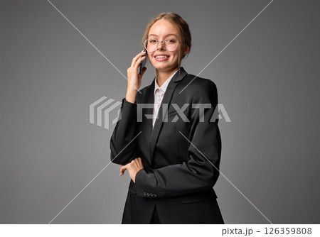 Cheerful businesswoman holding smartphone, smiling during phone call against grey background. Cheerful businesswoman holding smartphone, smiling during phone call against grey background. 126359808
