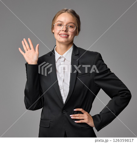 Smiling woman waving with hand in greeting, positive gesture, wearing formal black suit against grey studio background. 126359817