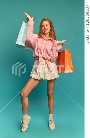 Happy young woman in pink shirt proudly showing shopping bags and smiling confidently against teal backdrop. 126359837