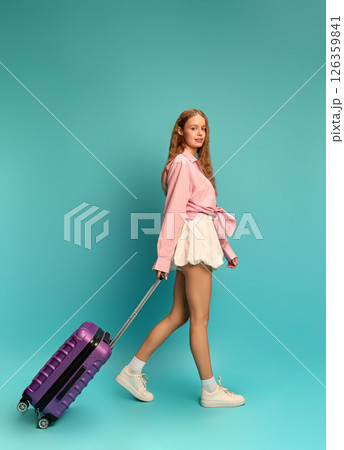 Young stylish woman in pink shirt and skirt walking with purple suitcase against teal backdrop. Vacation. Young stylish woman in pink shirt and skirt walking with purple suitcase against teal backdrop. Vacation. 126359841