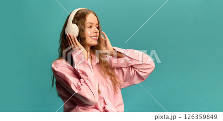 Young girl smiling and listening to music in wireless headphones, wearing pink shirt, posing on turquoise background. Young girl smiling and listening to music in wireless headphones, wearing pink shirt, posing on turquoise background. 126359849