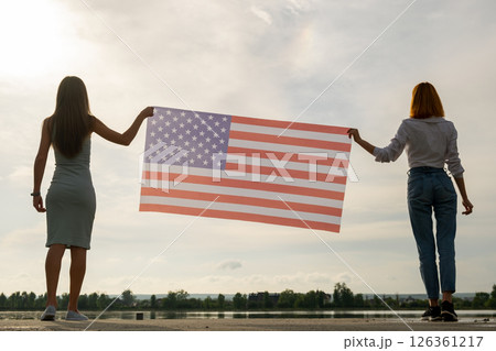 Silhouette of two young friends women holding USA national flag in their hands standing together outdoors. Patriotic girls celebrating United States independence day 126361217