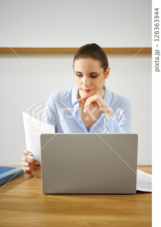 Focused woman reviewing documents while working on a laptop at a wooden desk in a bright office Focused woman reviewing documents while working on a laptop at a wooden desk in a bright office 126363944