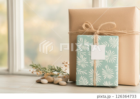 Two beautifully wrapped gifts with natural twine bows sit on wooden surface near smooth stones and greenery, evoking calm and warmth 126364733