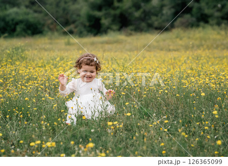 Beautiful happy little baby girl sitting on a green meadow with yellow flowers Beautiful happy little baby girl sitting on a green meadow with yellow flowers 126365099