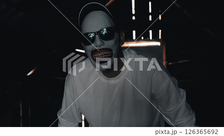 A man in a scary mask wearing a white sweatshirt and black cap is in a dimly lit attic with rays of light coming through the cracks. 126365692