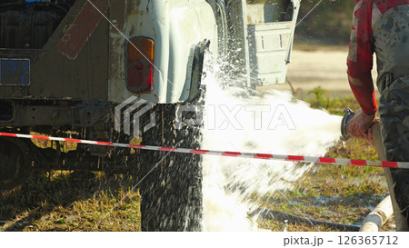 A person is washing a muddy off-road vehicle with a high-pressure hose. Water is spraying from the hose and hitting the vehicle with force, creating a cloud of spray. A person is washing a muddy off-road vehicle with a high-pressure hose. Water is spraying from the hose and hitting the vehicle with force, creating a cloud of spray. 126365712