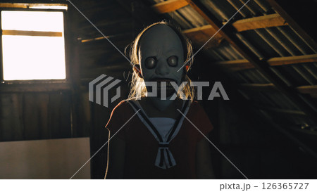 A young girl wearing a creepy mask with big eyes and a wide smile stands in a dark, shadow-covered attic. Her red sailor dress gives the scene a touch of disturbing innocence. A young girl wearing a creepy mask with big eyes and a wide smile stands in a dark, shadow-covered attic. Her red sailor dress gives the scene a touch of disturbing innocence. 126365727