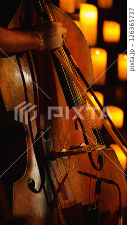 A close-up of a double bass being played in front of a wall of lit candles. The background is blurred and orange. 126365737