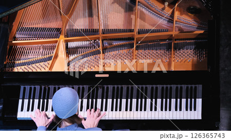 Top-down view of grand piano featuring copper-colored string frame and tuning pins above keyboard, with musician in blue jacket playing piano, hands spread across multiple octaves. Top-down view of grand piano featuring copper-colored string frame and tuning pins above keyboard, with musician in blue jacket playing piano, hands spread across multiple octaves. 126365743