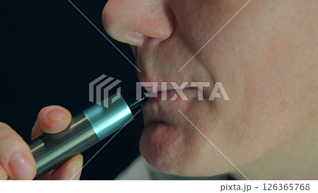 Person Using an Electronic Cigarette. A close-up of a person's face as they inhale from an electronic cigarette. The device is held in their hand. 126365768