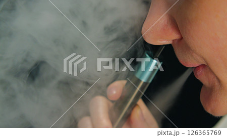 Close-up of a woman's face exhaling a cloud of vapor from an electronic cigarette. A hand holding a vape device, with thick clouds of vapor around it. Close-up of a woman's face exhaling a cloud of vapor from an electronic cigarette. A hand holding a vape device, with thick clouds of vapor around it. 126365769