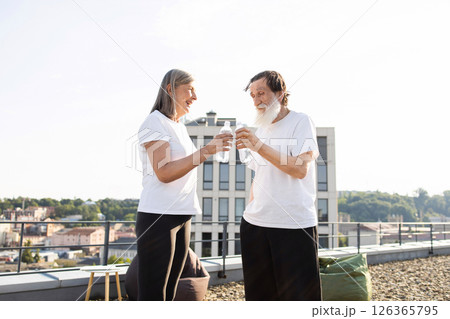 Elderly man and woman standing outside holding bottled water on rooftop after workout. Scene conveys feelings of health, companionship, and relaxation, with outdoor urban setting. 126365795
