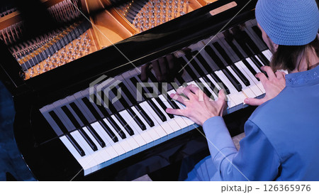 Pianist in blue clothes playing piano, hands resting on white keys. Naked copper-coloured piano strings and tuning pins. Top view. Pianist in blue clothes playing piano, hands resting on white keys. Naked copper-coloured piano strings and tuning pins. Top view. 126365976