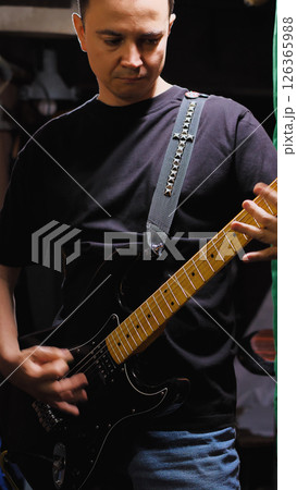 A person playing an electric guitar, wearing a black t-shirt and blue jeans. The guitar strap is decorated with metal studs. Guitarist at rehearsal point. 126365988