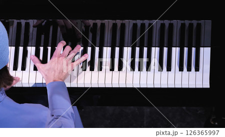Close-up view of pianist's hand stretched across black and white piano keys, wearing light blue sleeve, partial view of blue beanie visible, shot from above against dark background. 126365997