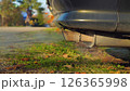 Close-up of a car's exhaust pipe emitting smoke. The car is parked on a grassy area with a sidewalk and trees in the background. Environmental Issues. Greenhouse gases. 126365998