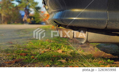 Close-up of a car's exhaust pipe emitting smoke. The car is parked on a grassy area with a sidewalk and trees in the background. Environmental Issues. Greenhouse gases. 126365998