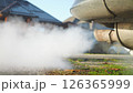 Close-up of the exhaust pipe of an old car with a lot of smoke coming out of it. The car is parked on a grassy area near a house. In background is a quiet street with houses. Environmental issues. 126365999