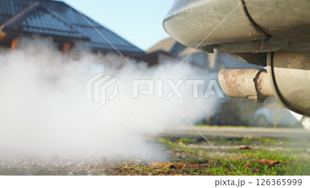 Close-up of the exhaust pipe of an old car with a lot of smoke coming out of it. The car is parked on a grassy area near a house. In background is a quiet street with houses. Environmental issues. Close-up of the exhaust pipe of an old car with a lot of smoke coming out of it. The car is parked on a grassy area near a house. In background is a quiet street with houses. Environmental issues. 126365999