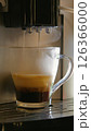 A close-up of a glass cup coffee machine pouring a cappuccino. Steam rises from the cup and the machine. A black espresso machine with a silver handle. The cup stands on a silver tray. 126366000