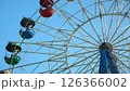 A low-angle shot of a Ferris wheel with colorful passenger cars against a clear blue sky. The intricate metal structure of the wheel is visible. 126366002