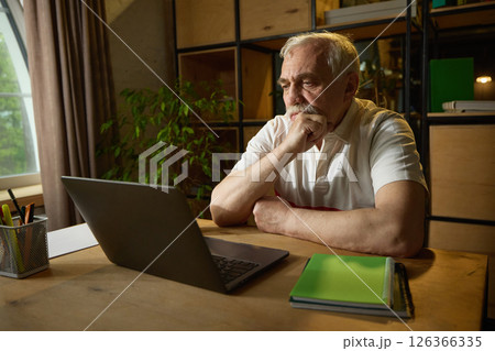 Elderly man with mustache sitting at wooden desk, looking thoughtfully at laptop with hand on chin, mug and notebooks nearby. Elderly man with mustache sitting at wooden desk, looking thoughtfully at laptop with hand on chin, mug and notebooks nearby. 126366335