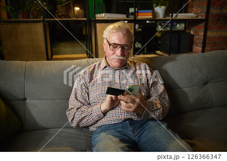 Elderly man sitting on grey sofa using smartphone and credit card for online shopping at home. Modern shopping activity 126366347