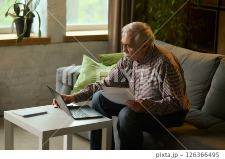 Focused elderly man sitting on sofa in modern living room, reading printed papers and using laptop for education, remote work. 126366495