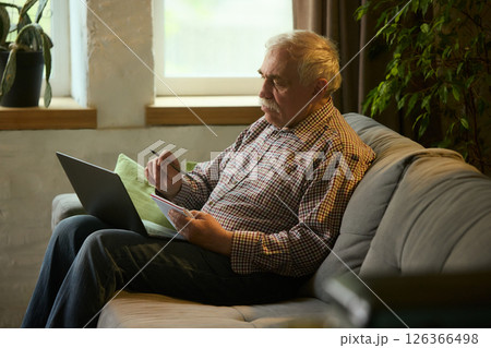 Senior man sitting on sofa at home in living room with laptop and notepad, taking notes during online class or research session. 126366498