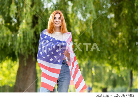 Happy young red haired woman posing with USA national flag standing outdoors in summer park. Positive girl with United States banner outdoors. 126367492
