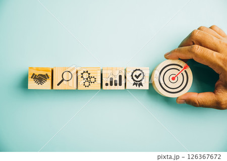 Hand arranging wood block stacking to illustrate the business strategy and action plan on a blue background. Showcasing the concept of business development and targeting success. Copy space available. Hand arranging wood block stacking to illustrate the business strategy and action plan on a blue background. Showcasing the concept of business development and targeting success. Copy space available. 126367672