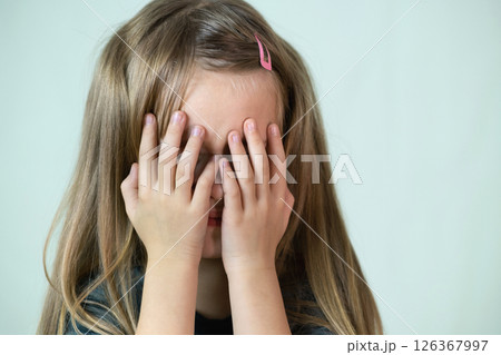 Close-up portrait of unhappy little girl with long hair covering her face with hands crying. 126367997