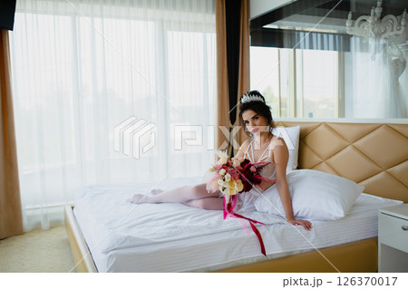 Seductive slender young bride in a sexy lingerie and white stockings posing on a bed with her bridal bouquet 126370017