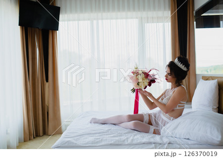 Seductive slender young bride in a sexy lingerie and white stockings posing on a bed with her bridal bouquet 126370019