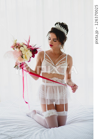 Seductive slender young bride in a sexy lingerie and white stockings posing on a bed with her bridal bouquet 126370025