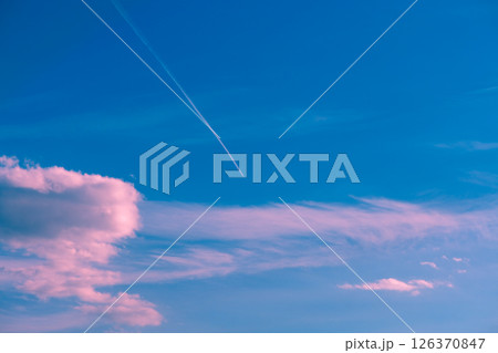 White contrail from a high-altitude airplane flying in a vibrant blue sky, with pink and white clouds adding a touch of color and serenity to the vast expanse 126370847