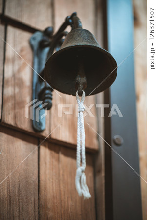 Hanging on a rustic wooden door, an old metal bell with a white rope awaits ringing, ready to announce arrivals or signal important events with its classic sound Hanging on a rustic wooden door, an old metal bell with a white rope awaits ringing, ready to announce arrivals or signal important events with its classic sound 126371507