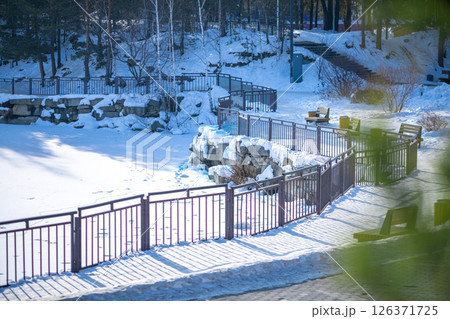 Winding metal fences encircle a frozen pond blanketed in snow, creating a serene winter park scene. Benches and trees enhance the tranquil atmosphere, inviting peaceful moments 126371725