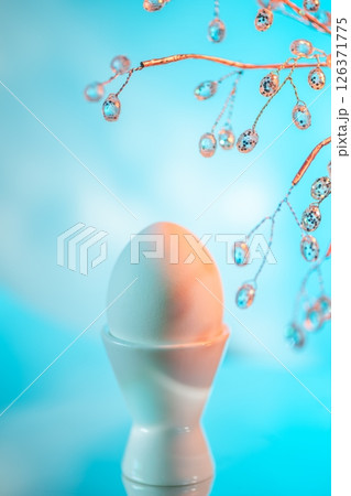 Easter egg in a white egg cup with the shadow of a decorative branch projected on it, creating a beautiful and festive atmosphere on a gradient blue background Easter egg in a white egg cup with the shadow of a decorative branch projected on it, creating a beautiful and festive atmosphere on a gradient blue background 126371775