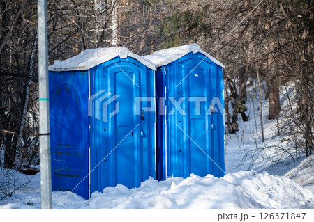Two blue portable toilet cabins are standing on a snowy ground in winter, surrounded by leafless trees and bushes, providing necessary facilities in outdoor environments 126371847