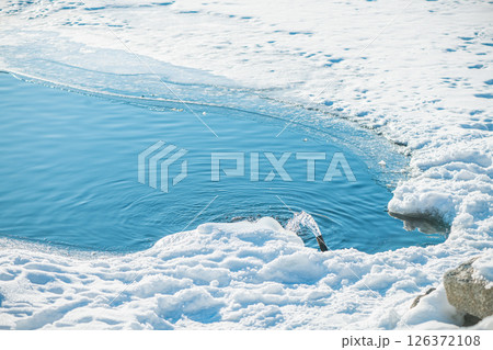 Water gushing from a pipe into a vibrant blue polynya amidst a snow-covered, icy landscape, creating a striking contrast between the cold environment and flowing water 126372108