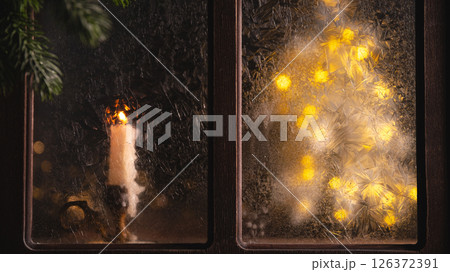 Burning candle sits on a windowsill next to a frosted window pane with a christmas tree made of string lights glowing behind it during a cold winter night Burning candle sits on a windowsill next to a frosted window pane with a christmas tree made of string lights glowing behind it during a cold winter night 126372391