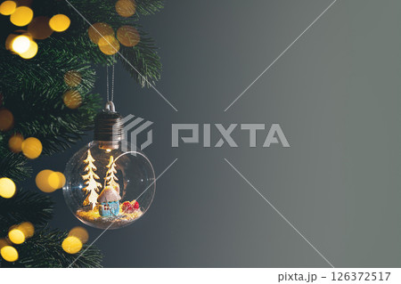 Glass light bulb ornament containing miniature snowy house and trees hanging on christmas tree branch against dark background. Copy space, greeting card 126372517