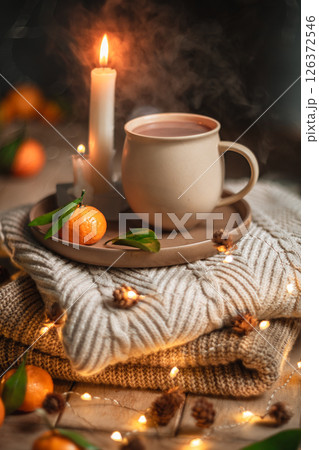 Steaming cup of hot beverage sits on a tray with a mandarin orange and lit candles, placed on a cozy knitted blanket with warm, inviting lighting, creating a perfect winter atmosphere Steaming cup of hot beverage sits on a tray with a mandarin orange and lit candles, placed on a cozy knitted blanket with warm, inviting lighting, creating a perfect winter atmosphere 126372546