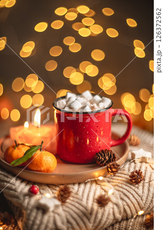 Red mug filled with hot chocolate and marshmallows resting on a tray with tangerines, surrounded by warm knit fabric and glowing christmas lights, creating a cozy winter scene 126372562